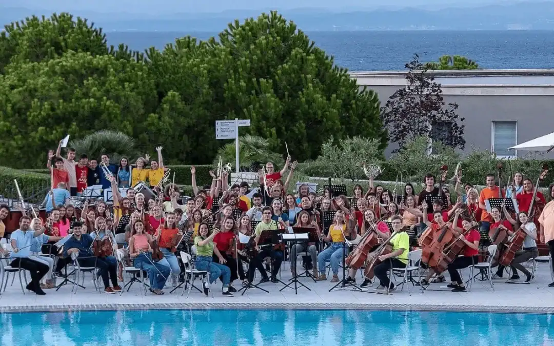 Vibrant youth orchestra performing outdoors near a swimming pool with lush greenery and ocean views in the background.