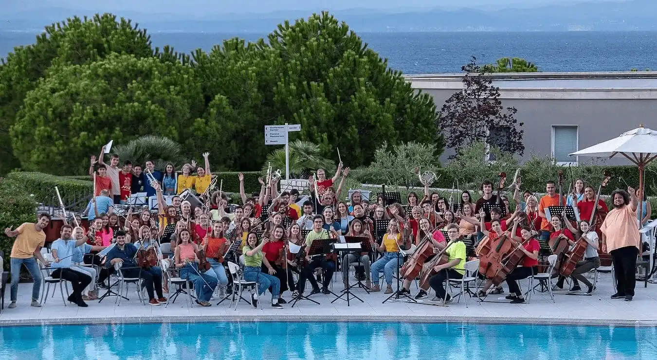 Vibrant youth orchestra performing outdoors near a swimming pool with lush greenery and ocean views in the background.